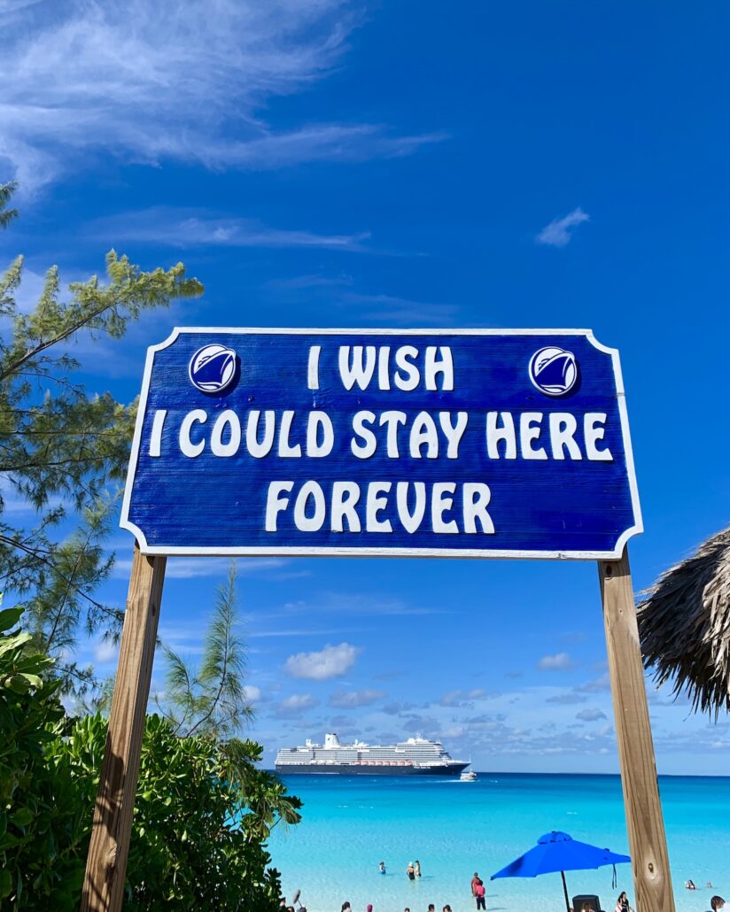 A blue sign reads "I wish I could stay here forever" with a beach, turquoise water, people swimming, a cruise ship in the distance, and a bright blue sky in the background.