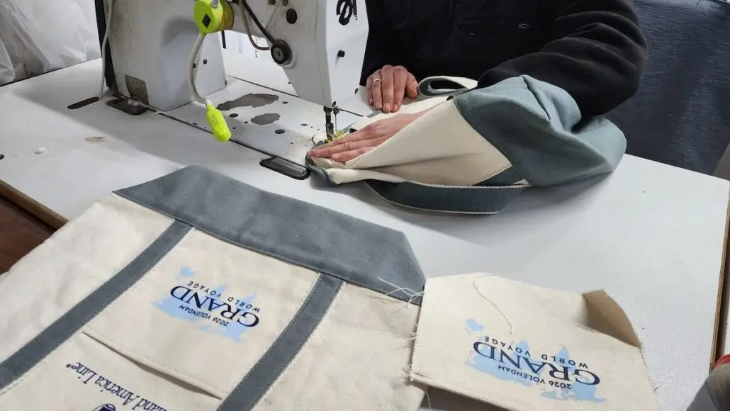 A person uses a sewing machine to stitch a fabric bag. Two completed tote bags and one unfinished piece, all labeled "GRAND WORLD VOYAGE 2024," lie on the table.