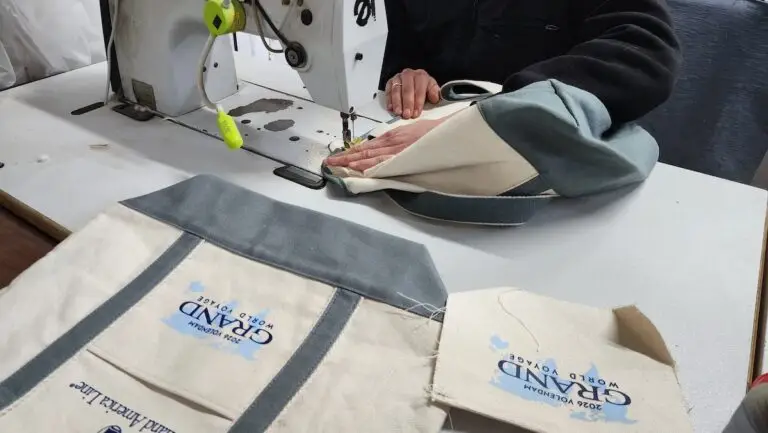 A person uses a sewing machine to stitch a fabric bag. Two completed tote bags and one unfinished piece, all labeled "GRAND WORLD VOYAGE 2024," lie on the table.