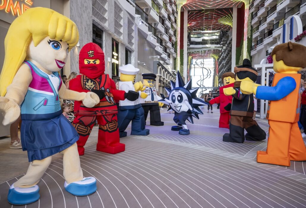 Several people dressed in large LEGO character costumes stand and interact in the lobby of a modern indoor venue with multiple floors and decorative lighting overhead.