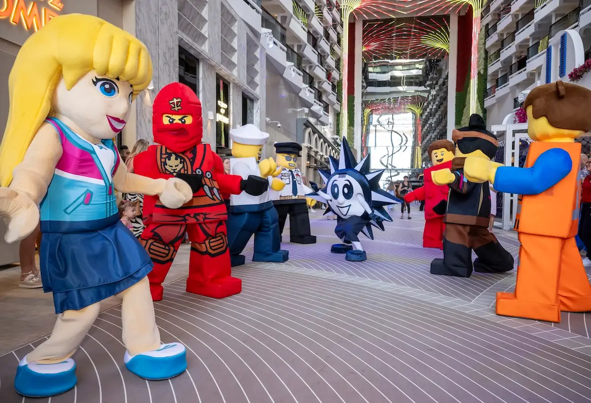 Several people dressed in large LEGO character costumes stand and interact in the lobby of a modern indoor venue with multiple floors and decorative lighting overhead.