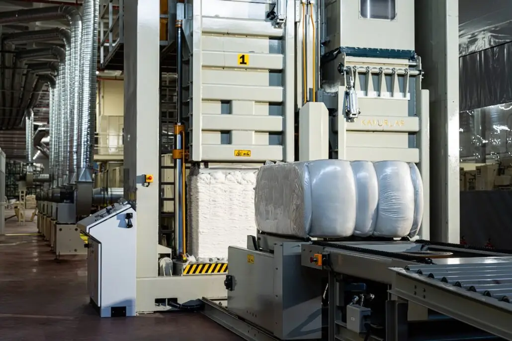 A factory interior shows large machines and wrapped bales of material on a conveyor belt, with industrial equipment, metal ducts, and a high ceiling visible in the background.