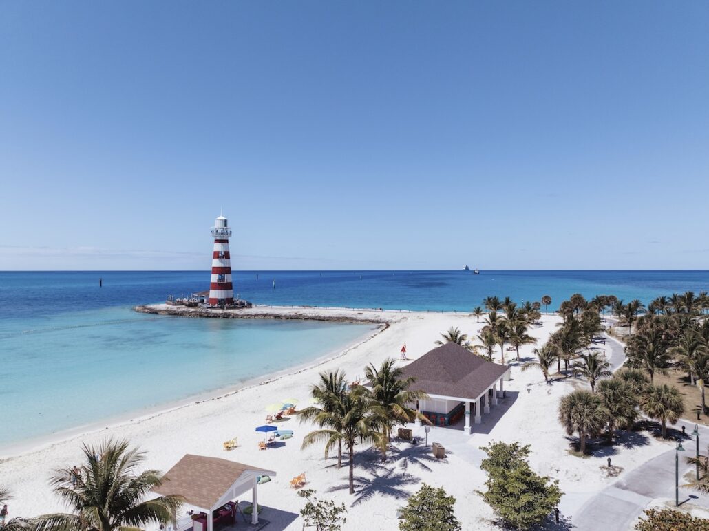 A red and white striped lighthouse stands on a small rocky peninsula surrounded by blue ocean water, with a sandy beach, palm trees, and several beach huts and chairs in the foreground under a clear sky.