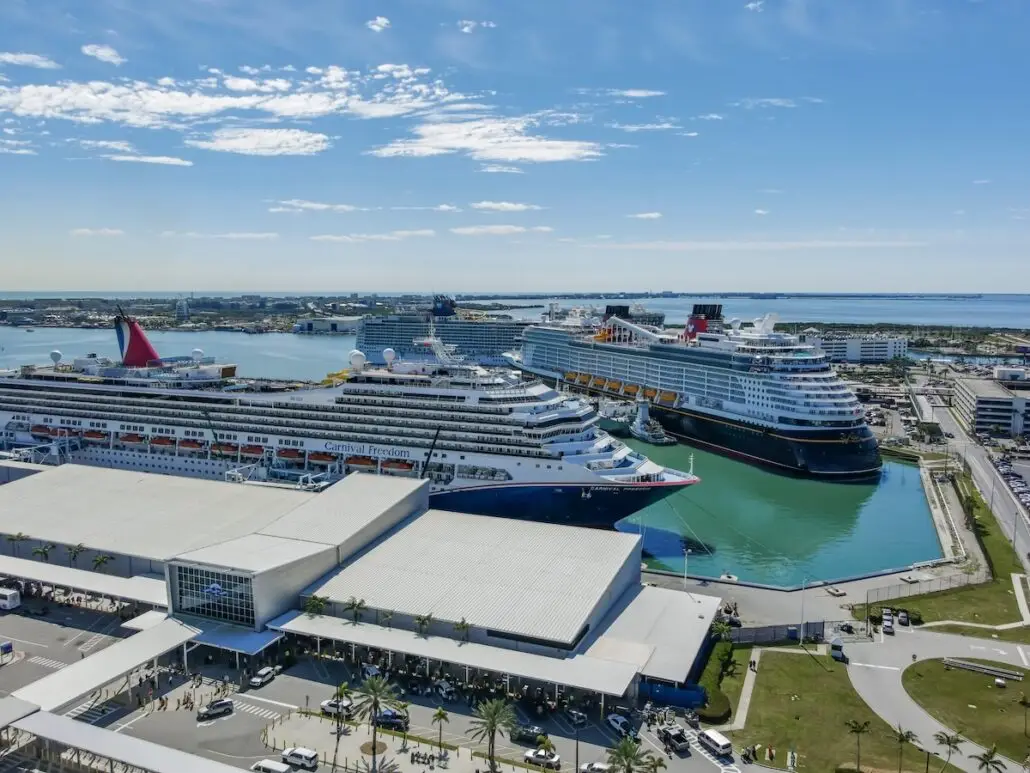 Aerial view of Port Canaveral, one of the busiest cruise ports, with multiple large cruise ships docked beside a terminal building, surrounded by parked vehicles and water under a partly cloudy sky.