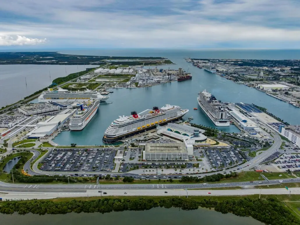Aerial view of Port Canaveral, the world's busiest cruise port in 2025, with several large cruise ships docked at multiple terminals, parking lots filled with cars, nearby buildings, and water extending inland under a partly cloudy sky.