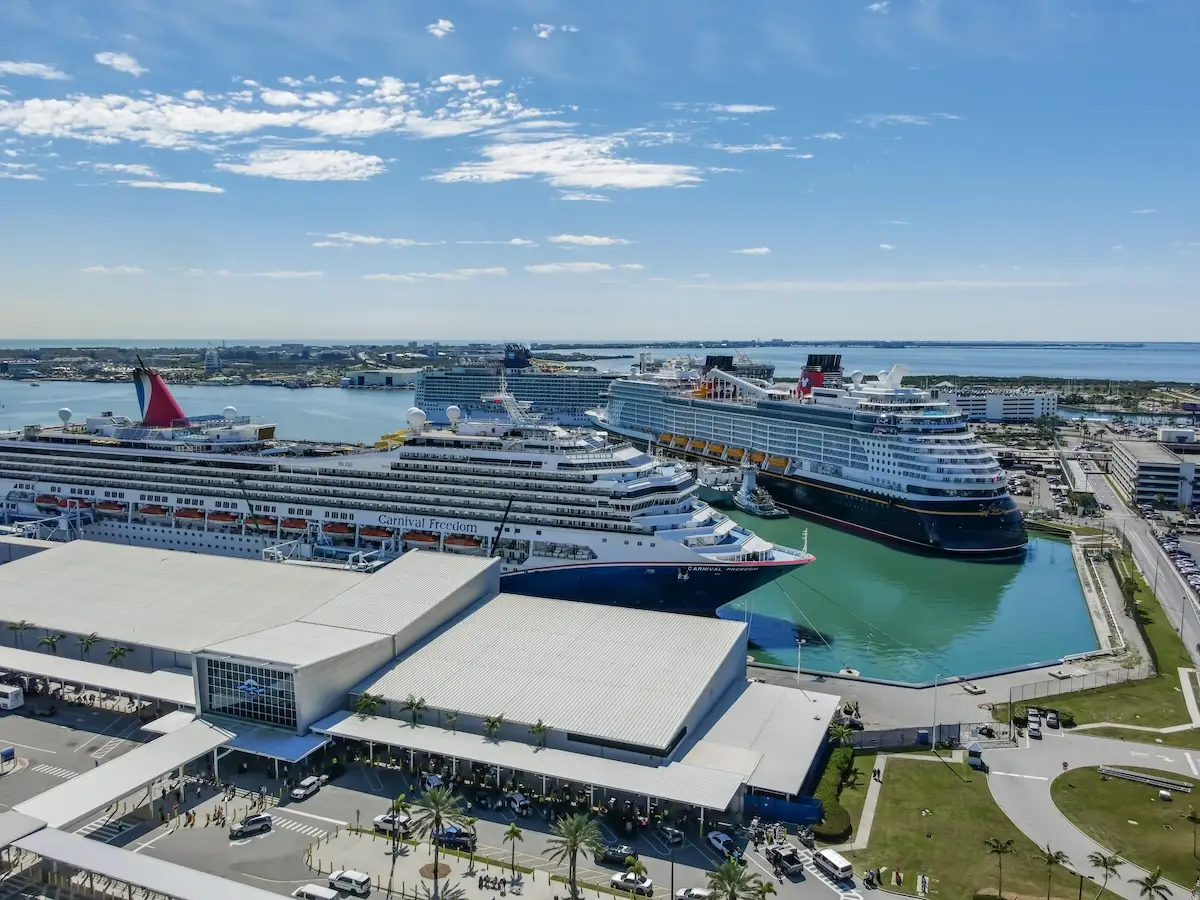 Aerial view of Port Canaveral, one of the busiest cruise ports, with multiple large cruise ships docked beside a terminal building, surrounded by parked vehicles and water under a partly cloudy sky.