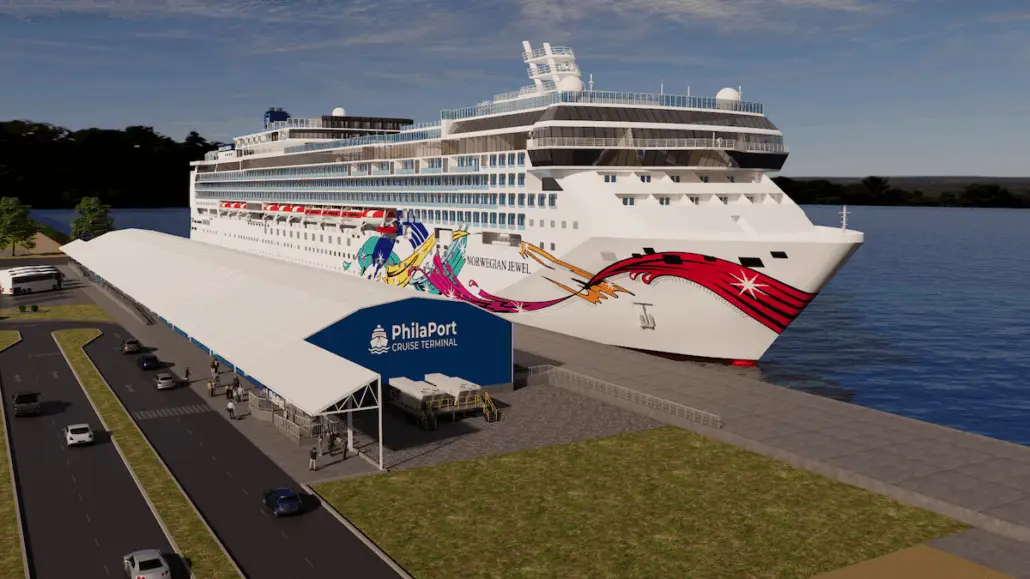 A large white cruise ship with colorful designs is docked at the PhilaPort Cruise Terminal. The terminal has a white roof and a blue sign. Several people and cars are visible near the terminal and along the road.