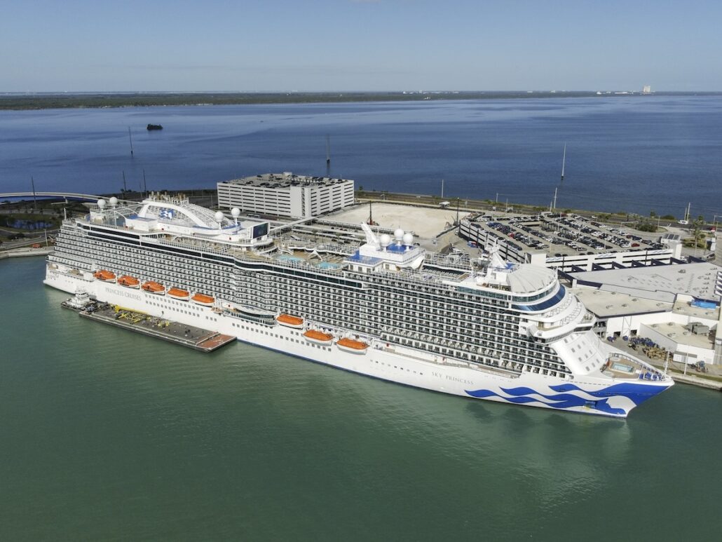 The Sky Princess, a large white Caribbean cruise ship, is docked at Port Canaveral near a parking garage, vehicles, and terminal buildings, all surrounded by calm blue water under a clear sky.