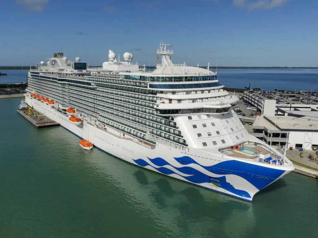 The Sky Princess, a large white cruise ship with blue wave designs, is docked at Port Canaveral beside a terminal building. Lifeboats line its side, with water and distant land visible under the clear sky—ready for its Winter Caribbean adventure.