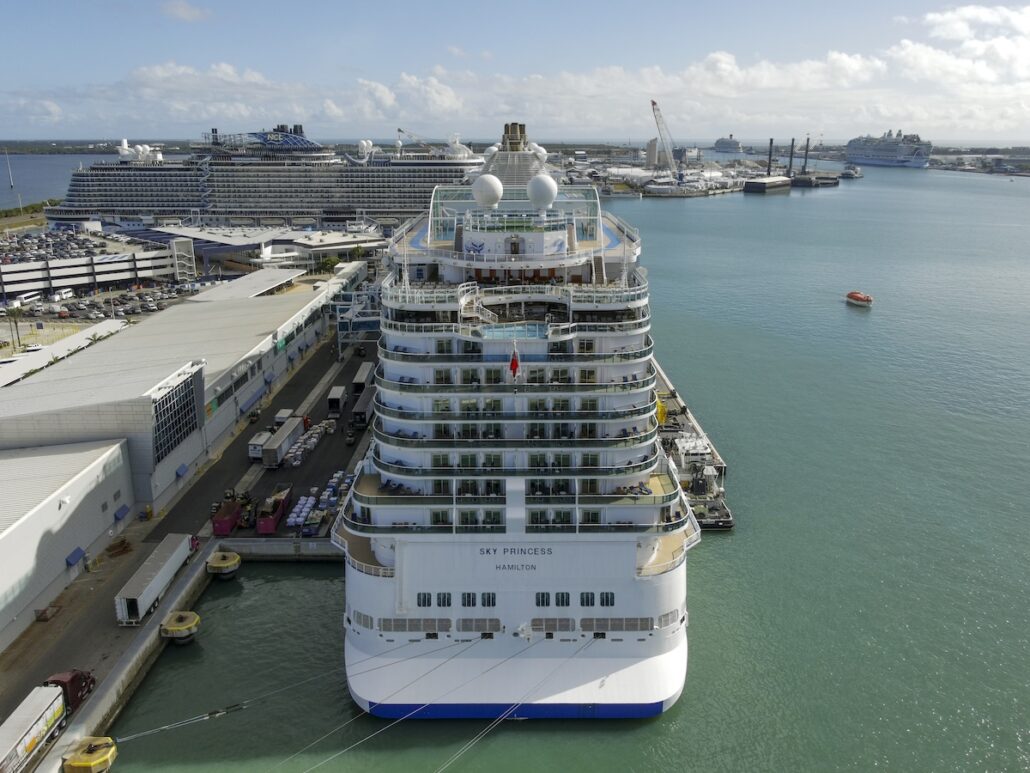 The Sky Princess, a large white cruise ship, is docked at Port Canaveral alongside several other vessels. The calm water reflects the busy port facilities and cranes in the background, hinting at upcoming Winter Caribbean adventures.
