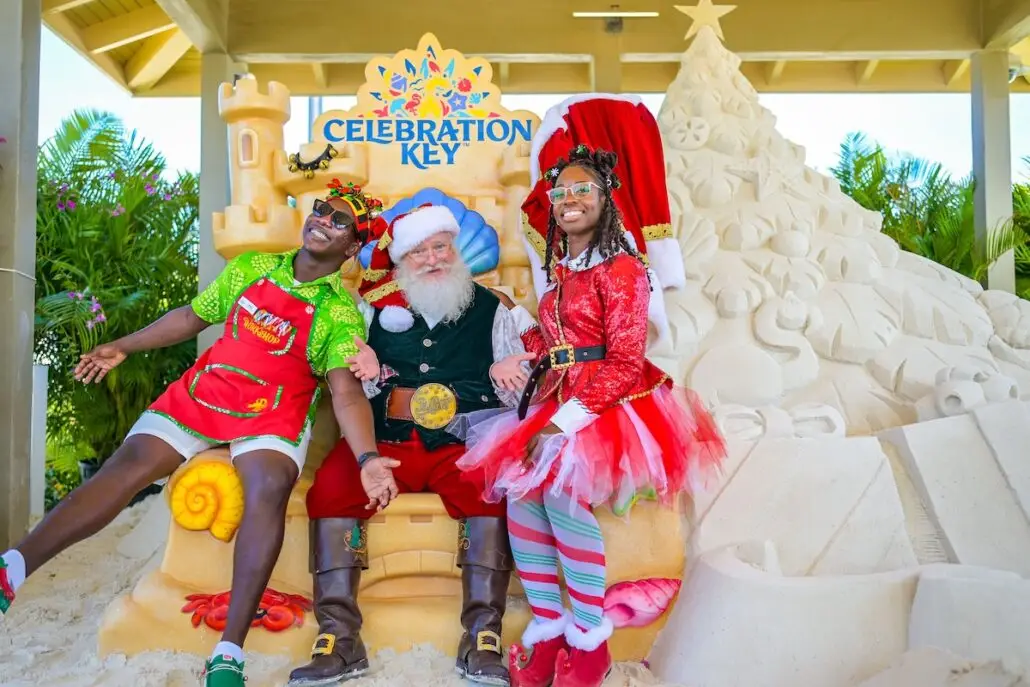 Three people pose in front of a sand sculpture with a “Celebration Key” sign. One person is dressed as Santa Claus, and the other two wear festive outfits. Palm trees and tropical plants are in the background.