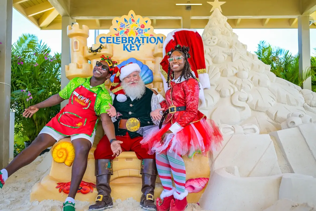 Three people pose in front of a sand sculpture with a “Celebration Key” sign. One person is dressed as Santa Claus, and the other two wear festive outfits. Palm trees and tropical plants are in the background.