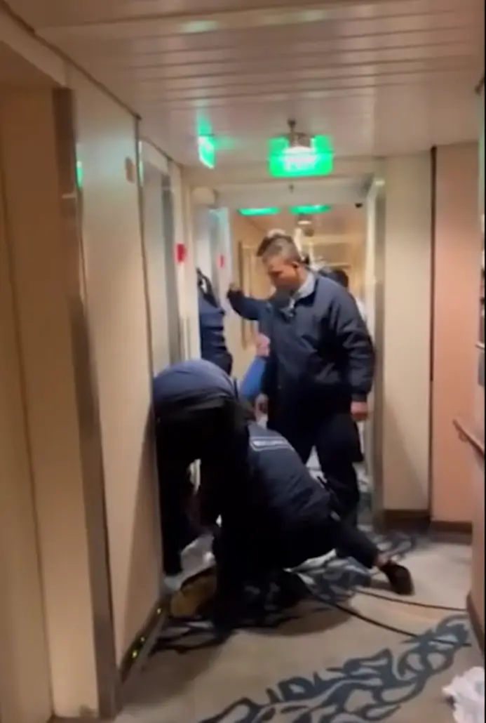 Three people in dark uniforms work in a narrow hallway with beige walls and patterned carpet. One kneels on the floor near a doorway, possibly investigating a guest death, while others stand observing. Overhead lights and green exit signs are visible.