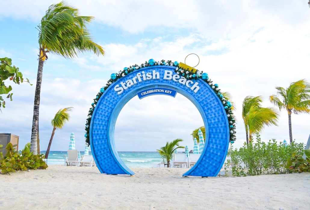 A large blue archway with the words "Starfish Beach" and "Celebration Key" stands on a sandy beach with palm trees, lounge chairs, and the ocean visible in the background.