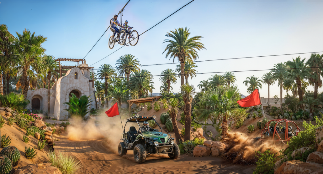 Two people ride bicycles on a high zipline above a sandy path near Ensenada’s cruise port, where an off-road vehicle drives, raising dust. Palm trees, desert plants, and a stone building are visible under a clear sky.