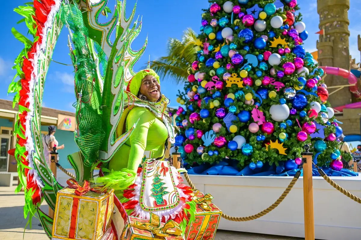 A performer in a bright green costume with feathered wings and gold-wrapped presents poses and smiles near a large, decorated Christmas tree outdoors under a sunny blue sky.
