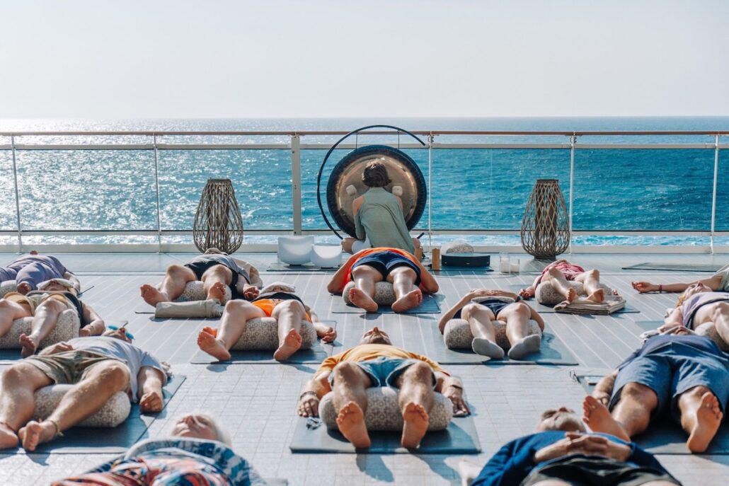 A group of people lie on yoga mats with their eyes closed on a deck overlooking the ocean, while an instructor at the front plays a large gong. The scene is bright and sunny with decorative lanterns on the railing.