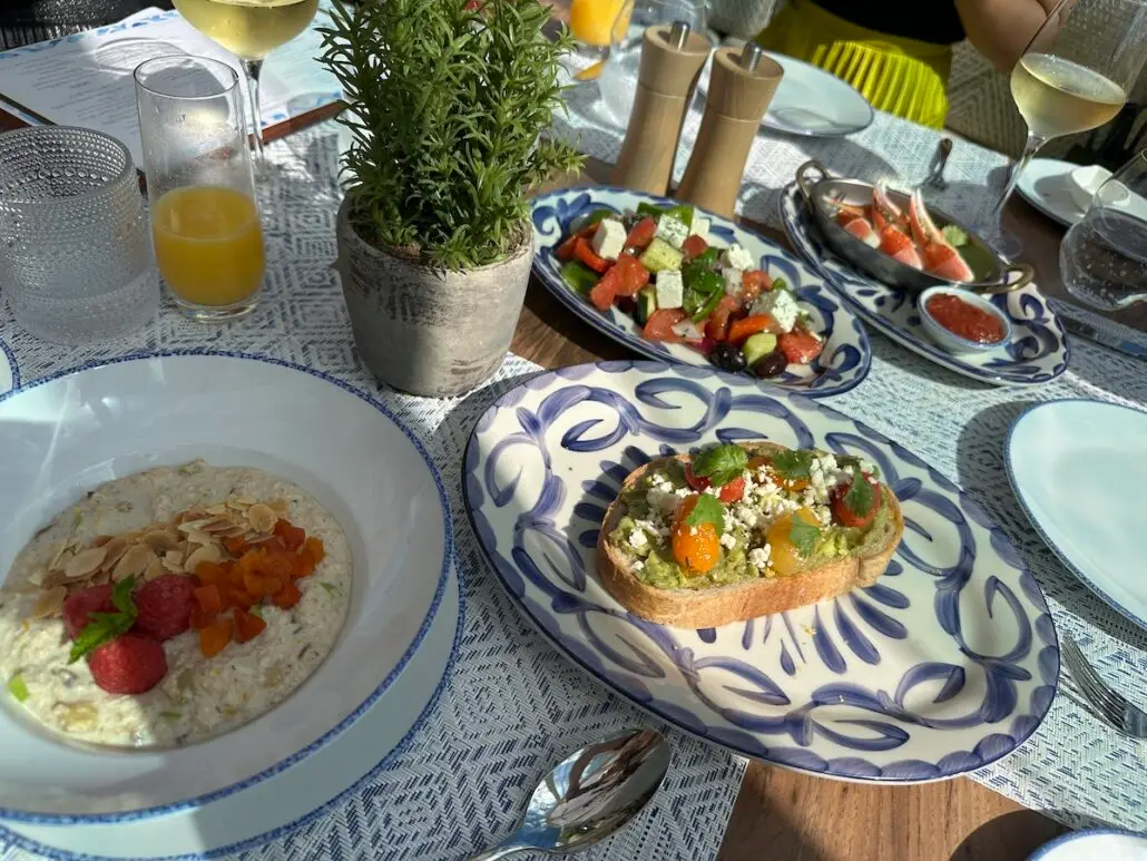 A table set for a meal aboard Celebrity Xcel features plates of food, including oatmeal with fruit and nuts, an open-faced sandwich with vegetables and cheese, Greek salad, shrimp cocktail, drinks, and a potted plant centerpiece.