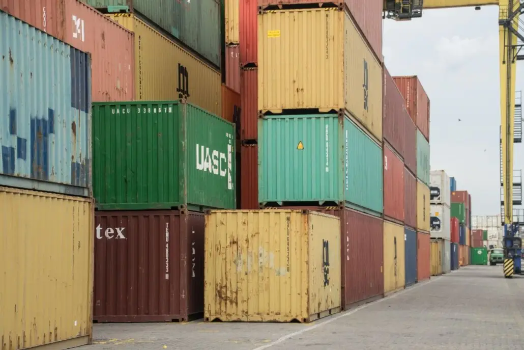 Stacks of colorful shipping and fruit containers are arranged in rows at a port, with a paved pathway alongside them. A crane structure is partially visible in the upper right corner, and a cloudy sky hints at a possible departure delay.