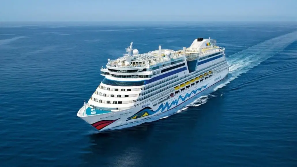 A large white cruise ship with colorful designs on the bow sails through calm blue ocean waters under a clear sky, as passengers remain vigilant about potential norovirus outbreaks during their fall illness season voyage.