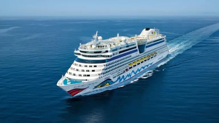 A large white cruise ship with colorful designs on the bow sails through calm blue ocean waters under a clear sky, as passengers remain vigilant about potential norovirus outbreaks during their fall illness season voyage.