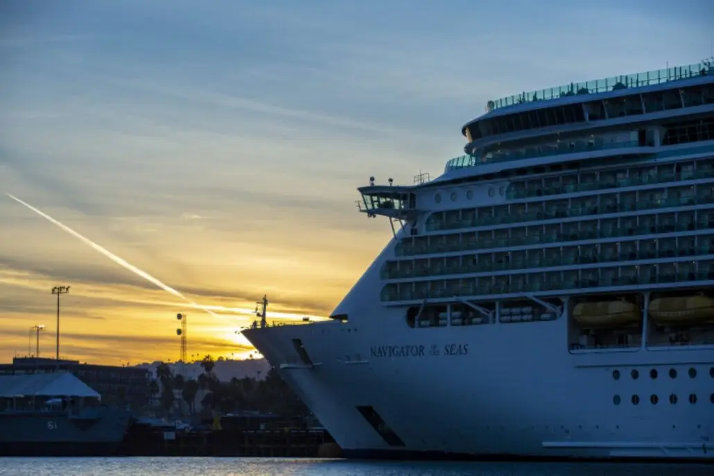 A large cruise ship named "Navigator of the Seas" docked at a port during sunset, with guests enjoying alcoholic drinks on deck, a contrail visible across the sky, and buildings silhouetted in the background.