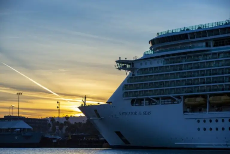 A large cruise ship named "Navigator of the Seas" docked at a port during sunset, with guests enjoying alcoholic drinks on deck, a contrail visible across the sky, and buildings silhouetted in the background.
