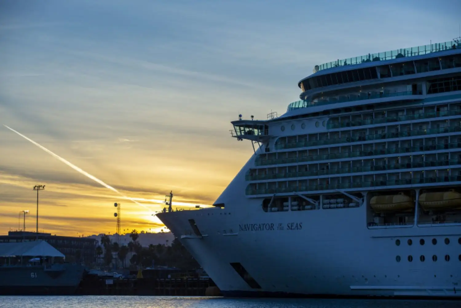 A large cruise ship named "Navigator of the Seas" docked at a port during sunset, with guests enjoying alcoholic drinks on deck, a contrail visible across the sky, and buildings silhouetted in the background.