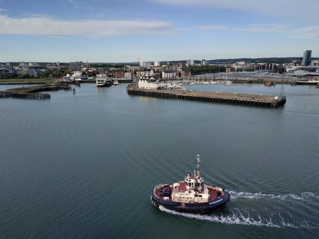 A small tugboat moves across calm water in a harbor, leaving a wake behind it. The harbor is lined with docks, fruit containers, and boats, with a cityscape and cloudy sky in the background.