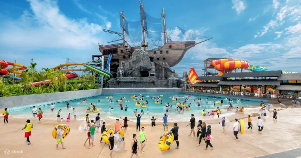 A large outdoor water park with many people in swimwear, a wave pool, inflatable rings, colorful water slides, and a large pirate ship structure under a bright blue sky.
