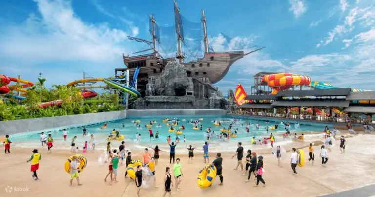 A large outdoor water park with many people in swimwear, a wave pool, inflatable rings, colorful water slides, and a large pirate ship structure under a bright blue sky.