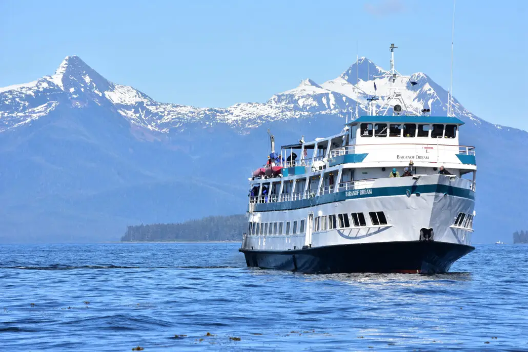 A white and blue ferry named "Baranof Dream," part of a local cruise line, travels on calm water with snow-capped mountains and a forested shoreline in the background under a clear blue sky.