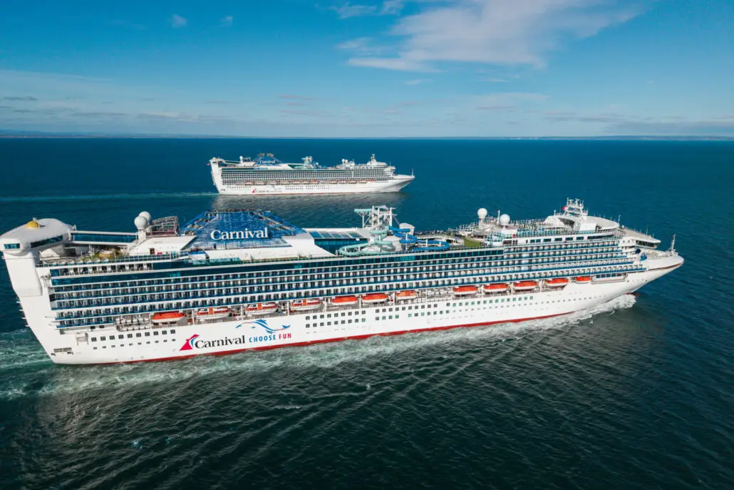 Two large Carnival cruise ships sail on calm blue water under a clear sky, moving parallel to each other. These Australia ships feature multiple decks with balconies, including a steakhouse and sushi restaurant onboard.