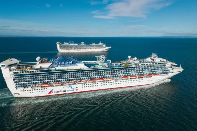 Two large Carnival cruise ships sail on calm blue water under a clear sky, moving parallel to each other. These Australia ships feature multiple decks with balconies, including a steakhouse and sushi restaurant onboard.