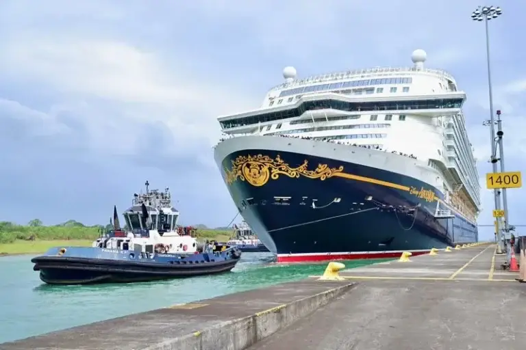 A large cruise ship, possibly the Disney Adventure or another of the largest ships, is assisted by a tugboat as it navigates through the Panama Canal beside a concrete dock with yellow safety barriers and signage under a cloudy sky.
