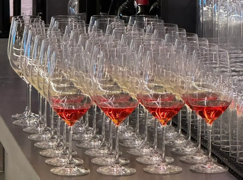 Rows of empty wine glasses are lined up on a counter, with the front row containing glasses partially filled with a red liquid—a setup reminiscent of one of John Heald’s iconic tasting events. Glasses and bottles can be seen in the background.