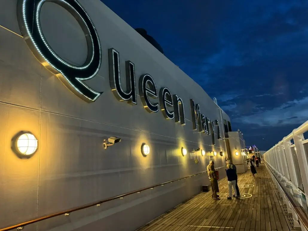 A nighttime view of the upper deck of a ship with illuminated "Queen Mary 2" signage, people walking along the wooden deck, and round lights lining the side—far from any epstein files cruise, just pure ocean elegance.