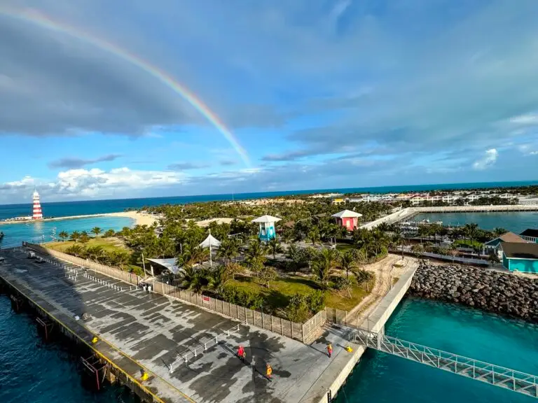 Aerial view of Ocean Cay, MSC Cruises’ private island, with a pier, turquoise water, a small lighthouse, palm trees, and colorful buildings; a rainbow arcs across a partly cloudy sky.
