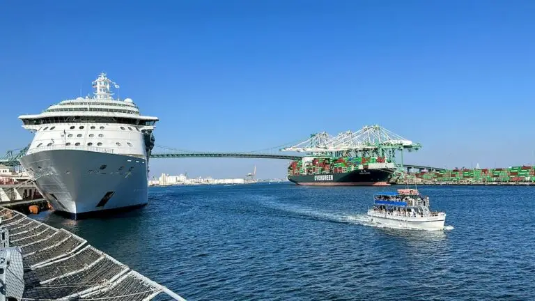 A large cruise ship is docked on the left at the Port of Los Angeles, a cargo ship loaded with containers is near cranes in the background, and a small boat is traveling on the water under a clear blue sky.