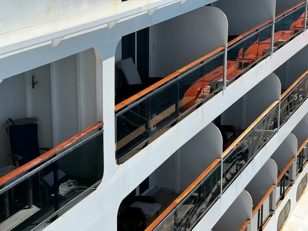 Close-up view of cruise line balconies with glass and wood railings. Orange lifeboats are visible below. Some balconies have chairs and a table, and part of the ocean can be seen in the background.
