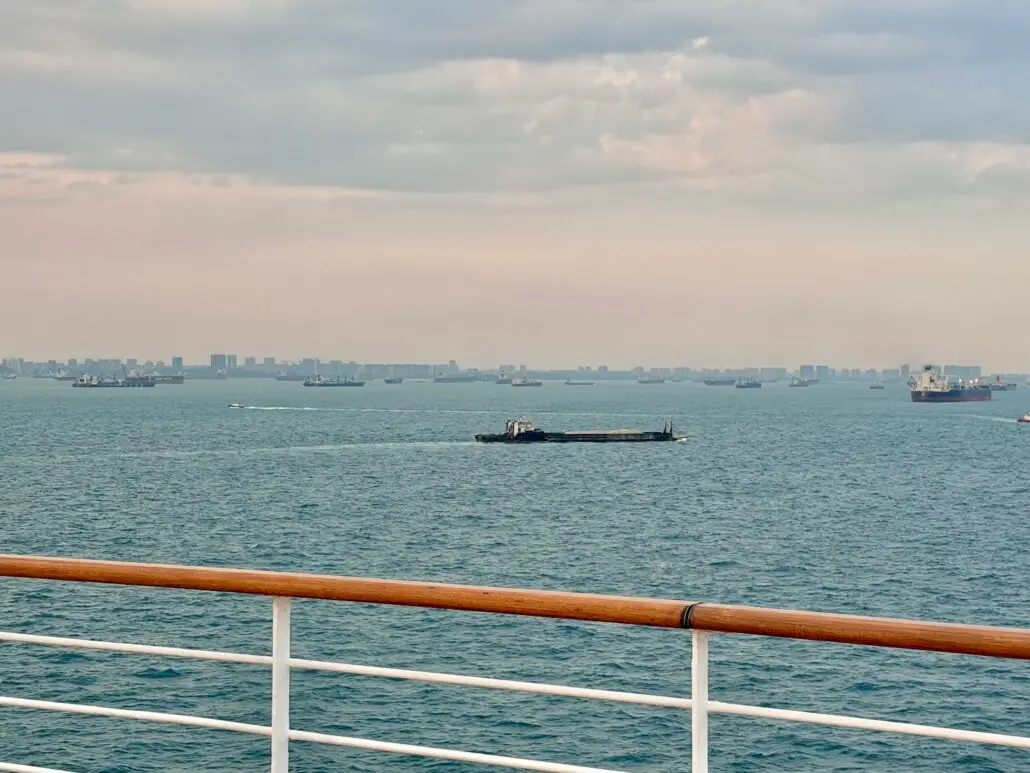 View from a ship deck with a wooden railing, overlooking several cargo ships and boats on a calm sea beneath a cloudy sky. The distant city skyline hints at Asia—a serene scene aboard a Holland America World Cruise.