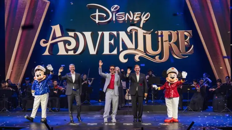 Four people, including two in Mickey and Minnie Mouse costumes, stand on stage waving in front of a large "Disney Adventure" sign, celebrating the launch of the Disney Cruise Line's newest ship, with an orchestra seated behind them.