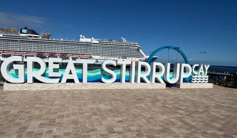 Large white letters spell out "Great Stirrup Cay" on a paved area of this private island, with a cruise ship docked in the background and a blue arch to the right, welcoming future guests under a clear sunny sky.