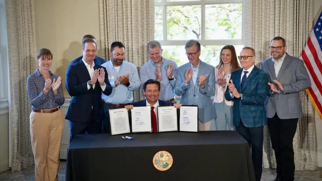 A group of people in business attire stand and applaud behind a seated man holding up two signed documents at a table with the Florida state seal. An American flag is visible on the right.