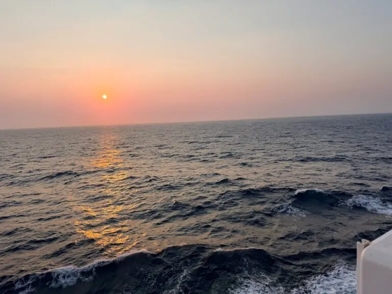 The sun is setting over a calm ocean with gentle waves, painting the perfect end to a day of travel on a world cruise. The sky is mostly clear with a slight haze, and part of a white structure is visible in the lower right corner.