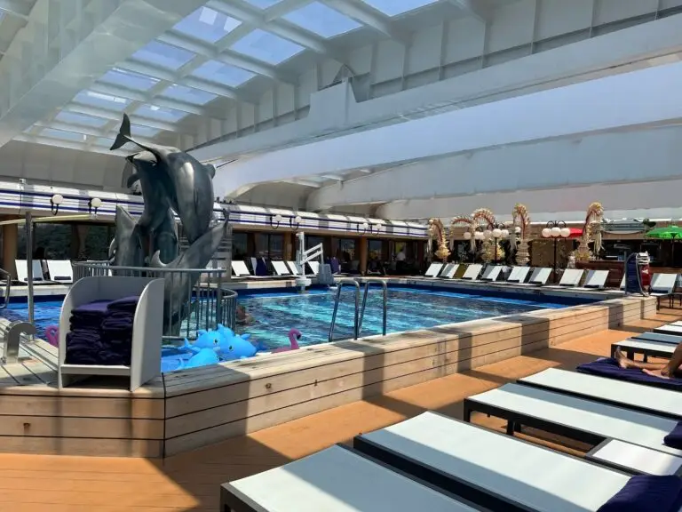 Indoor pool area with lounge chairs, a metal dolphin sculpture, and poolside decorations including inflatable toys—perfect for relaxing like you’re on a world cruise. The pool has railing access and sits beneath a glass ceiling letting in natural light.