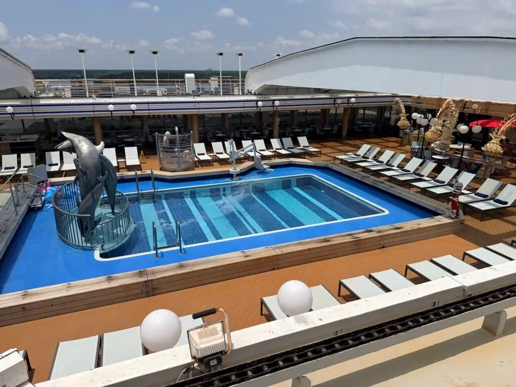 A rectangular swimming pool on a world cruise ship deck is surrounded by lounge chairs. A dolphin statue and water slide are at one end of the pool, with the partly cloudy sky and sea visible in the background.