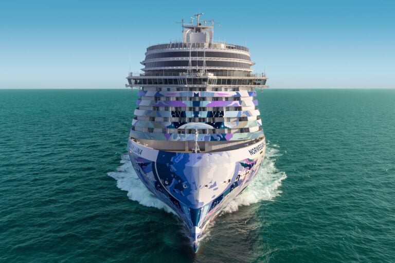 A large cruise ship is shown from the front, sailing through calm blue-green water under a clear sky. The ship features blue and purple artwork on its hull and has multiple decks visible.