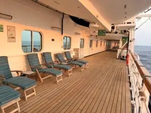 A row of striped lounge chairs sits on the wooden deck of a cruise ship, perfect for relaxing during a world cruise. The empty deck features safety signs, a railing to the right, and the ocean stretching into the distance.
