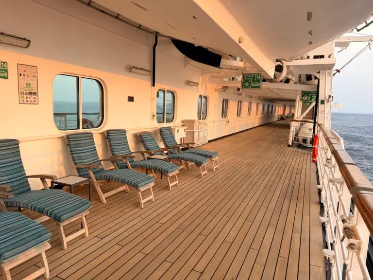 A row of striped lounge chairs sits on the wooden deck of a cruise ship, perfect for relaxing during a world cruise. The empty deck features safety signs, a railing to the right, and the ocean stretching into the distance.